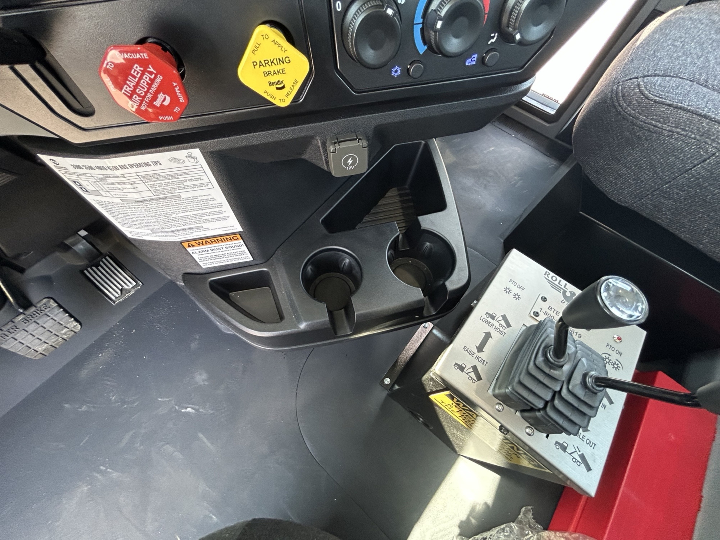 Cab interior of 2026 Mack GR64B rollover truck showing gear shifter panel, two cup holders, parking brake lever, and control switches with labeled decals.