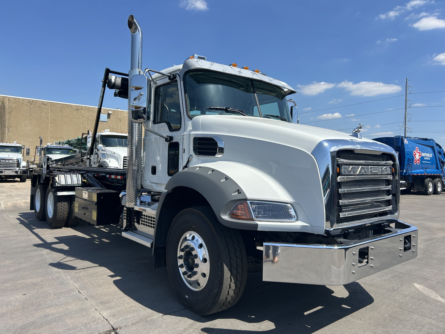 Truck: 2026 Mack GR64B Roll-off, front-left view showing chrome bumper, dual exhaust stack, large grille with Mack emblem, white cab, heavy-duty fenders, and attached trailer with metal decking.