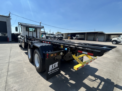 Mack GR64B roll-off truck chassis with flatbed and yellow-striped rear bumper, viewed from rear-right side. Includes black chassis, dual rear axles, mud flaps, and warning decals.