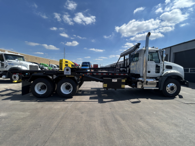Mack GR64B roll-off truck side view showing long flatbed, dual rear axles, chrome exhaust stack, and cab with gray paint.