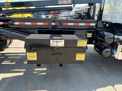 Grille-side toolbox mounted on a Mack GR64B rolloff truck showing black metal toolbox with yellow warning labels and mounted undercarriage equipment.