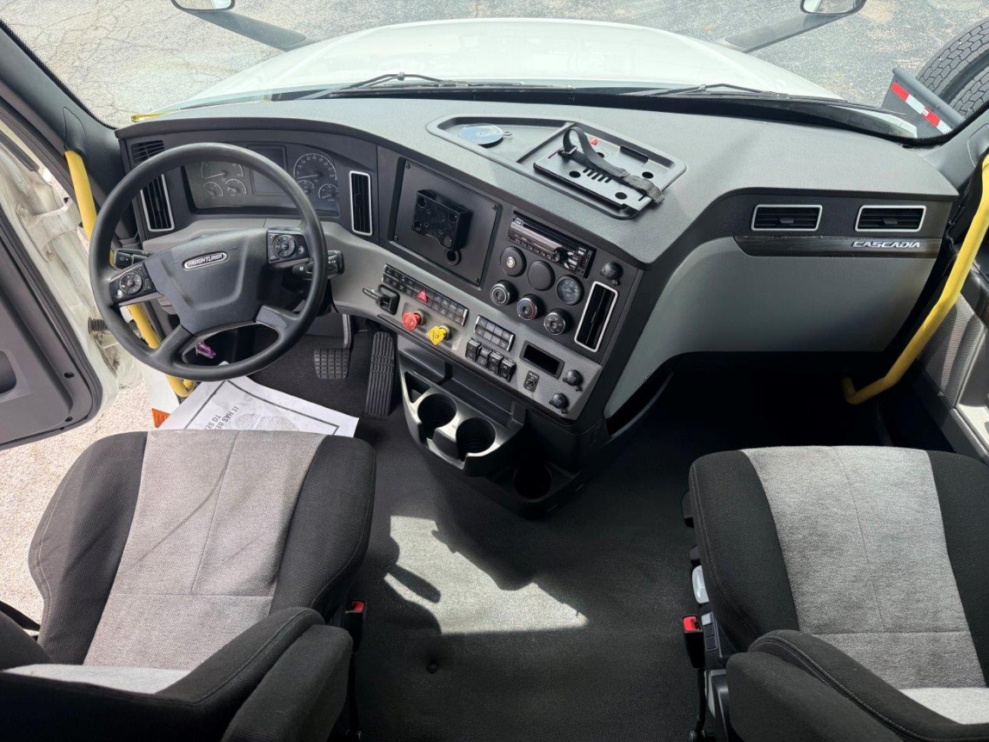 Truck cab interior showing steering wheel, dashboard with gauges, center console, and seating. Visible text includes Freightliner-branded steering wheel and console controls.