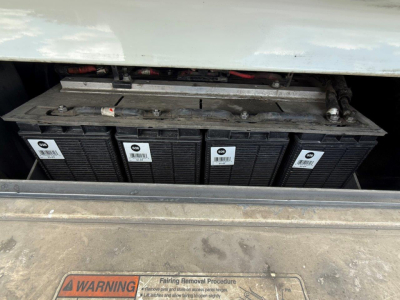 Truck battery bank in engine bay with multiple lead-acid cells and warning label visible; part of Freightliner Cascadia 126 under hood.