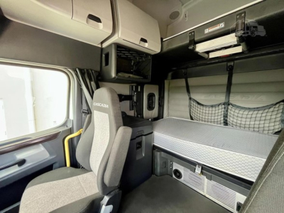 Cab interior of a Freightliner Cascadia 126 sleeper with bunk bed, storage compartments, and a gray driver seat; overhead luggage rack and controls visible.