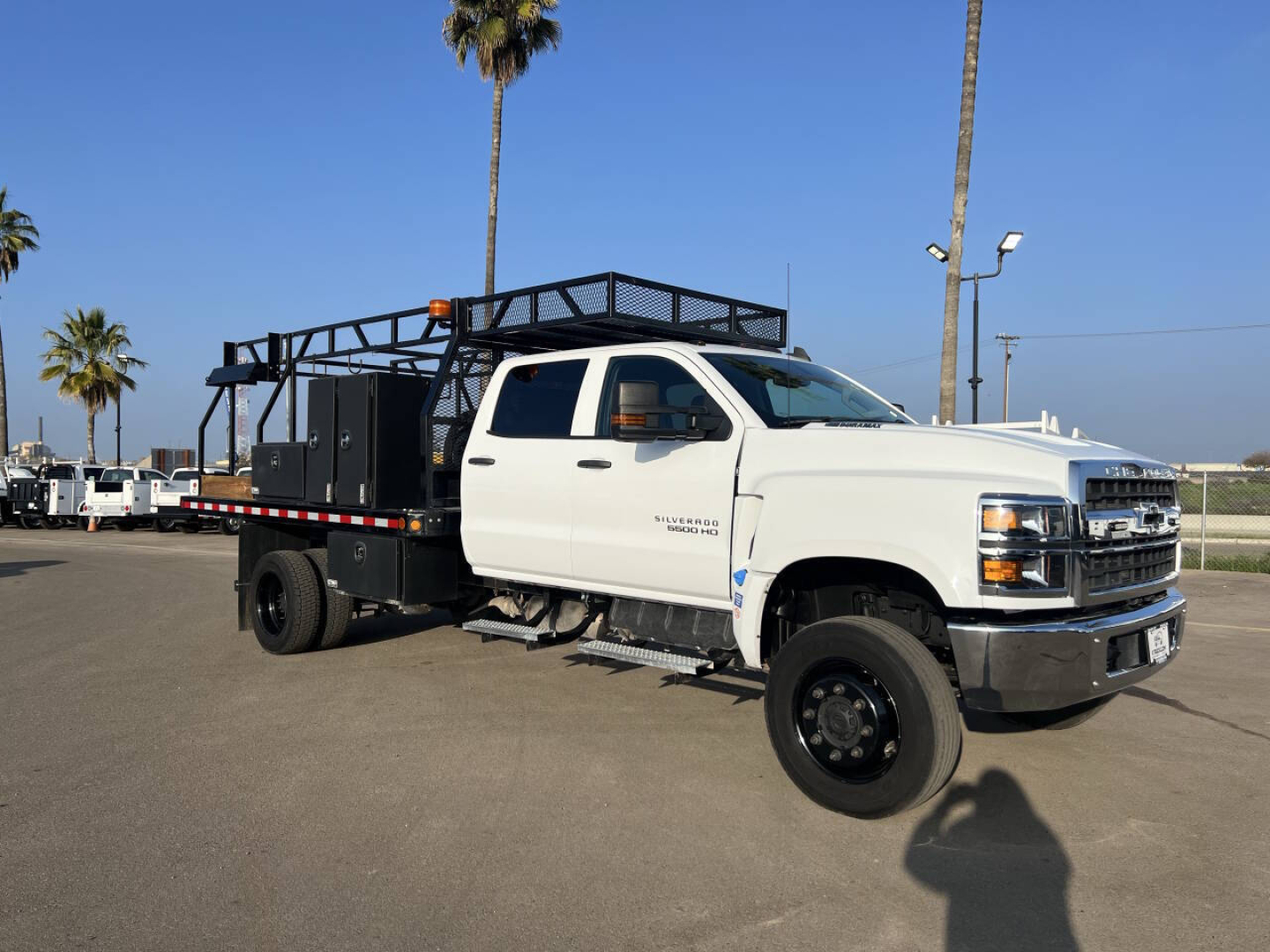 White Chevrolet Silverado flatbed tow truck with equipment racks and utility cage, showing a side view of the cab and long flatbed in a parking lot.