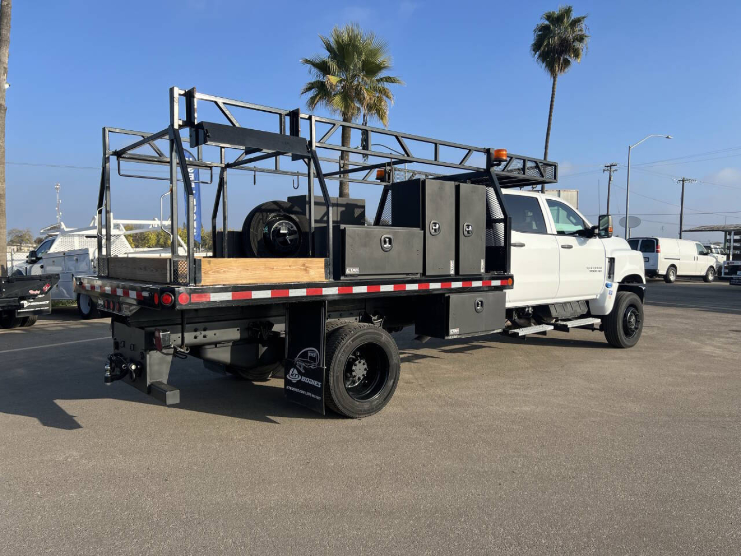Truck flatbed showing service compartments and rack with spare tire, viewed from left-rear side; white cab and black flatbed with toolboxes and ladder rack.