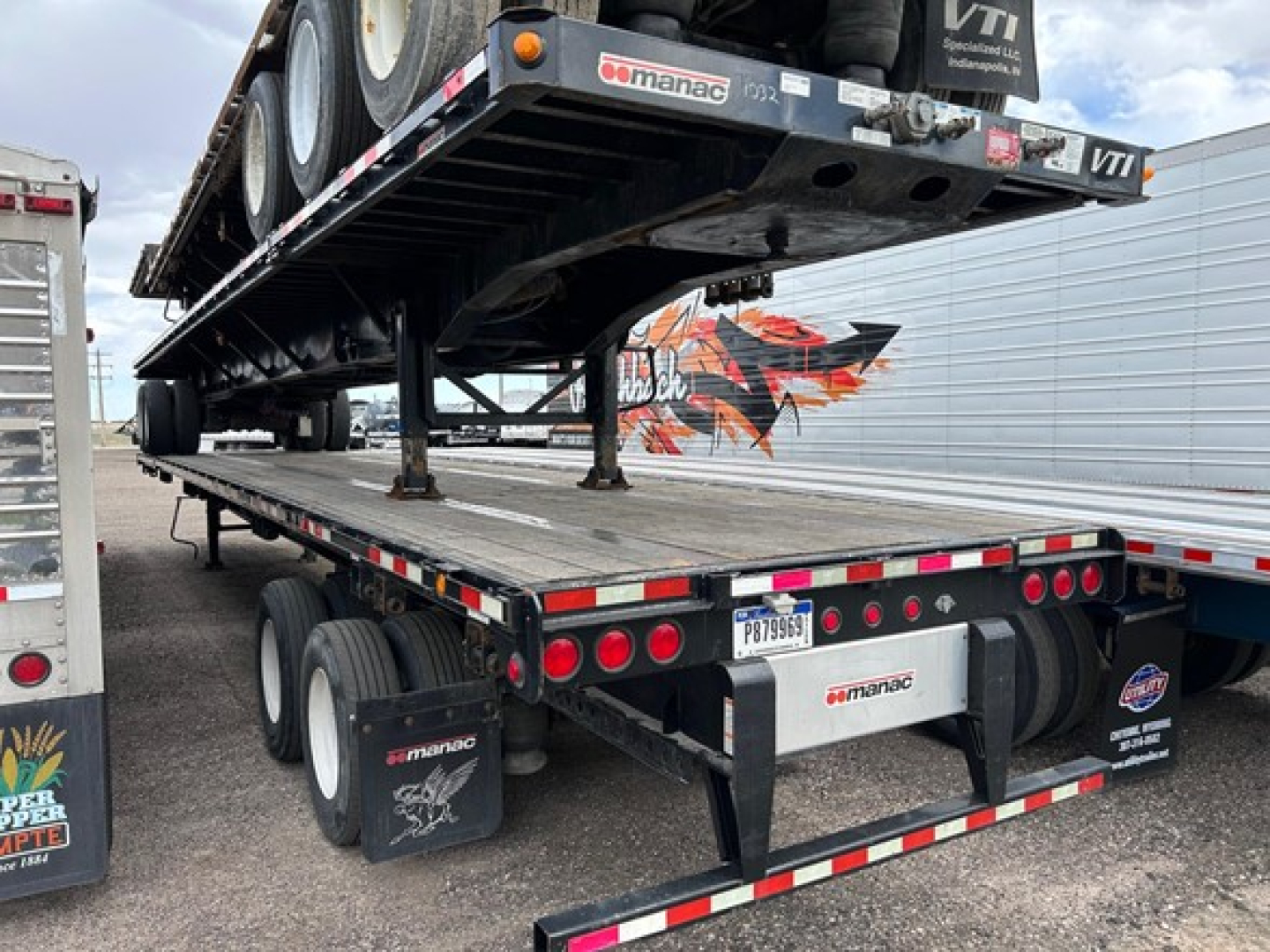 Extendable steel flatbed trailer with ramp and dual rear axles showing Manac branding and reflective tape.