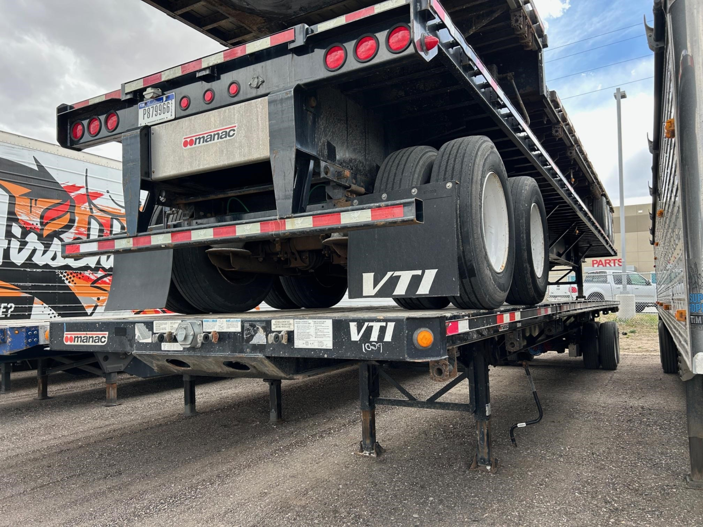 Flatbed trailer rear view showing extendable steel flatbed and wheels. Visible text includes Manac logo and VTI on the undercarriage and safety reflectors.