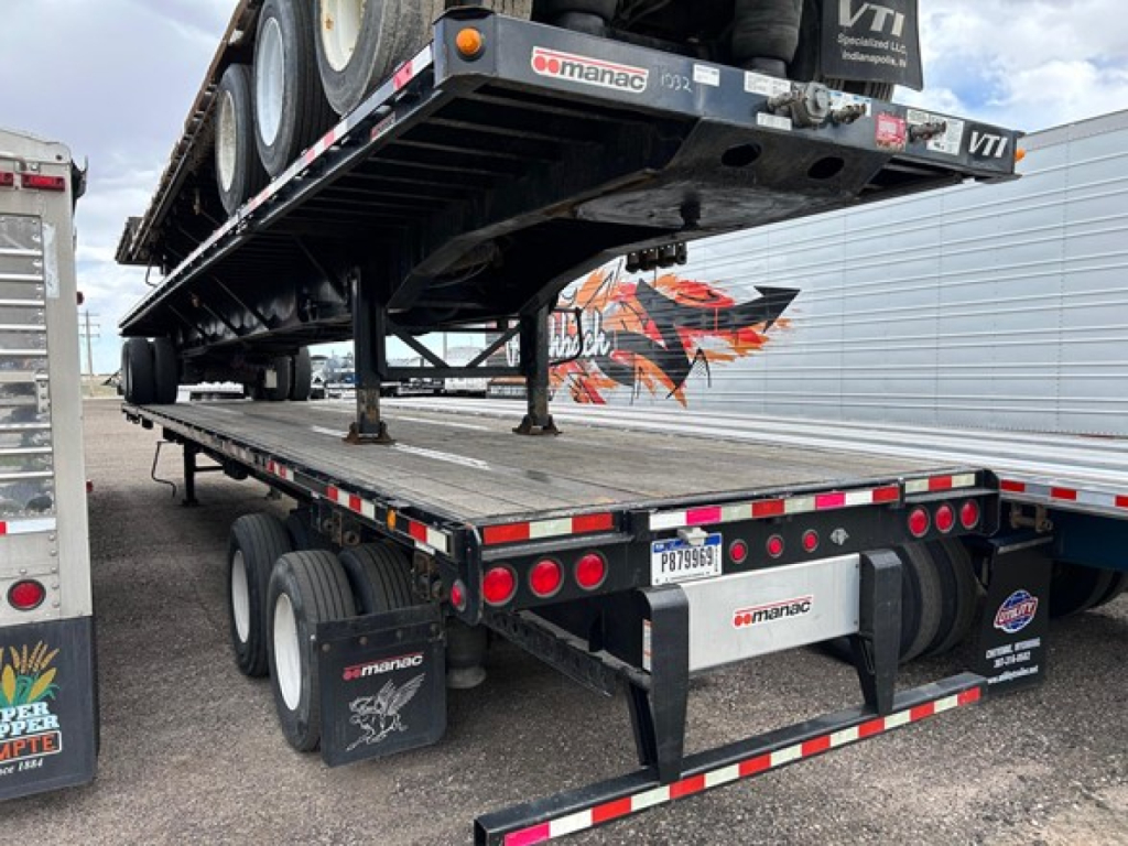 Extendable steel flatbed trailer with ramp and dual rear axles showing Manac branding and reflective tape.