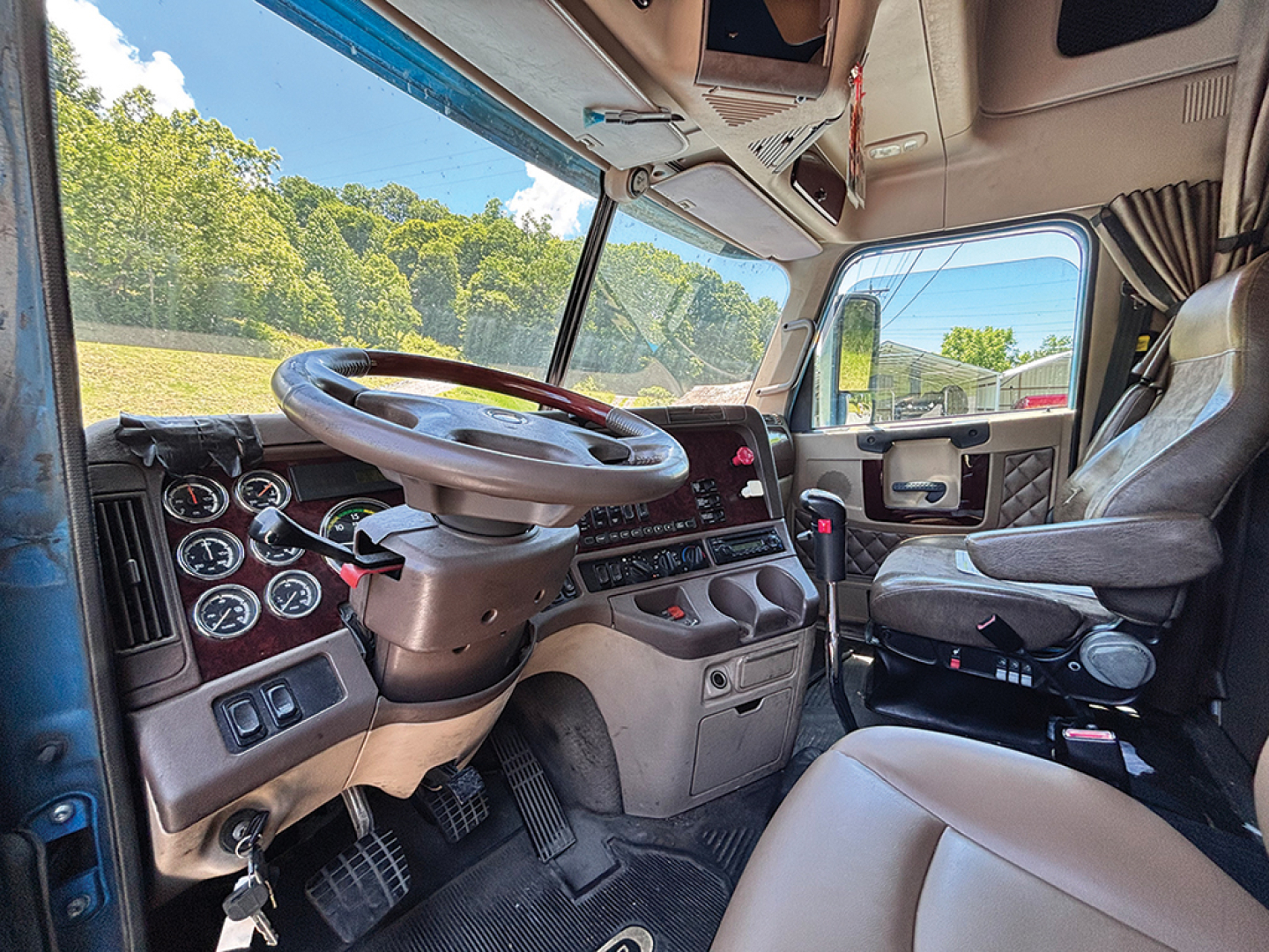 Cab view of a 2019 Kenworth W-900 AeroCab showing steering wheel, gauge cluster, and driver's seat. Text on dashboard and controls visible.