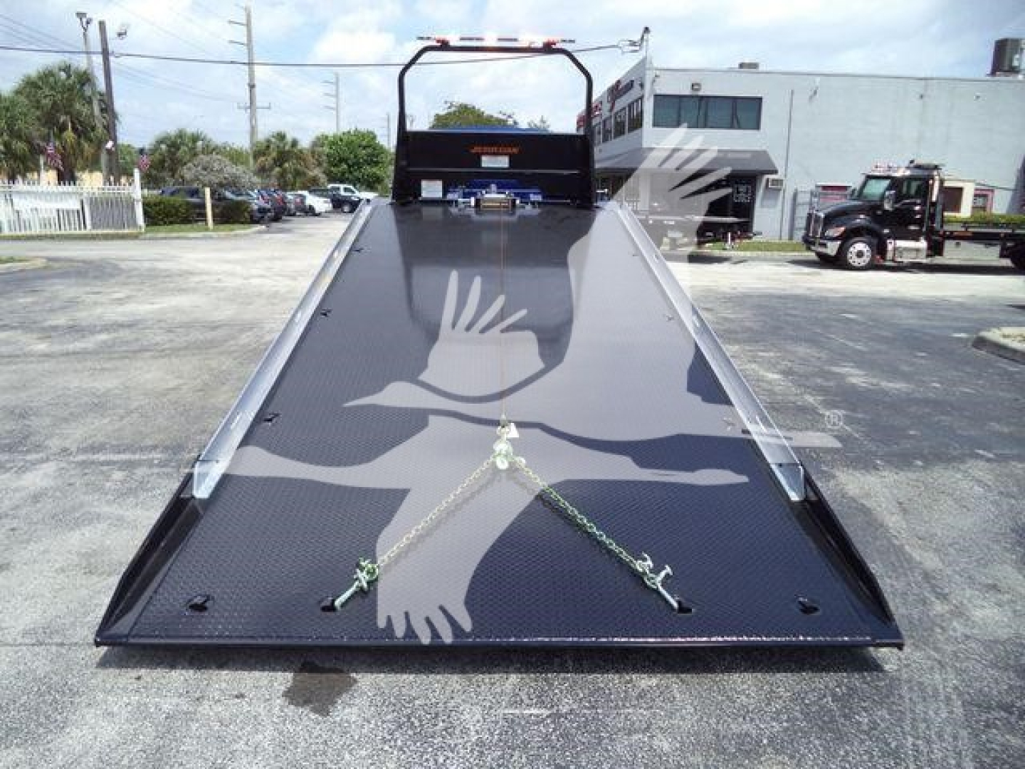 Flatbed tow truck ramp showing black steel surface and tie-down chains, with a rear cockpit bench and hitch in the distance;MV607EXCABBLU brand tag not readable.