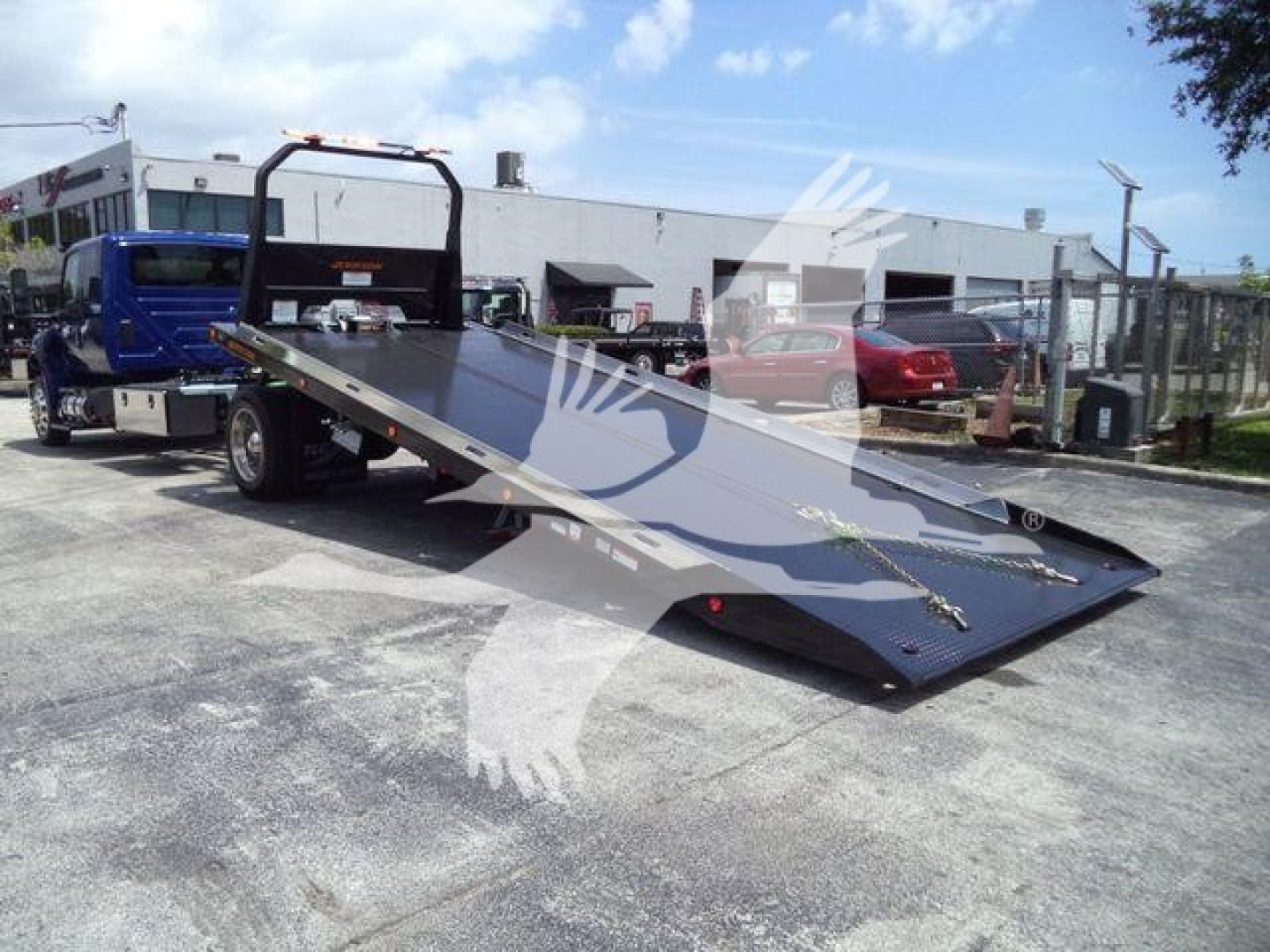 Flatbed tow truck with a long black metal ramp at an outdoor lot; surrounding vehicles and a building in background.