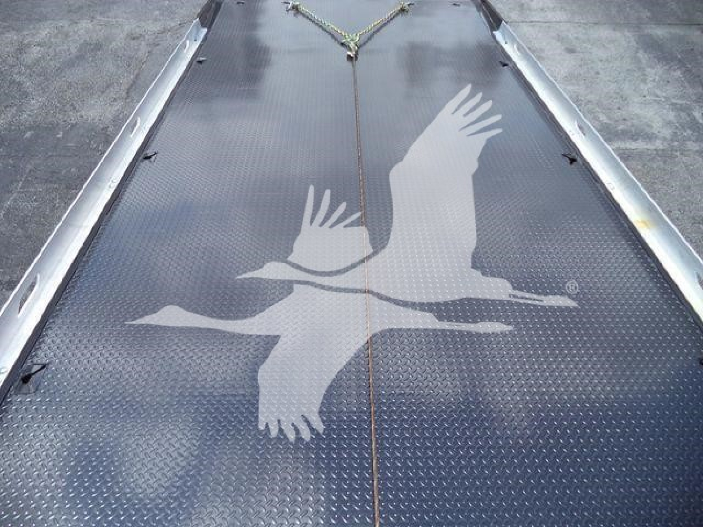 Flatbed truck deck with diamond plate surface featuring a large white flying geese graphic in the center and tied down metal rails along the edges.