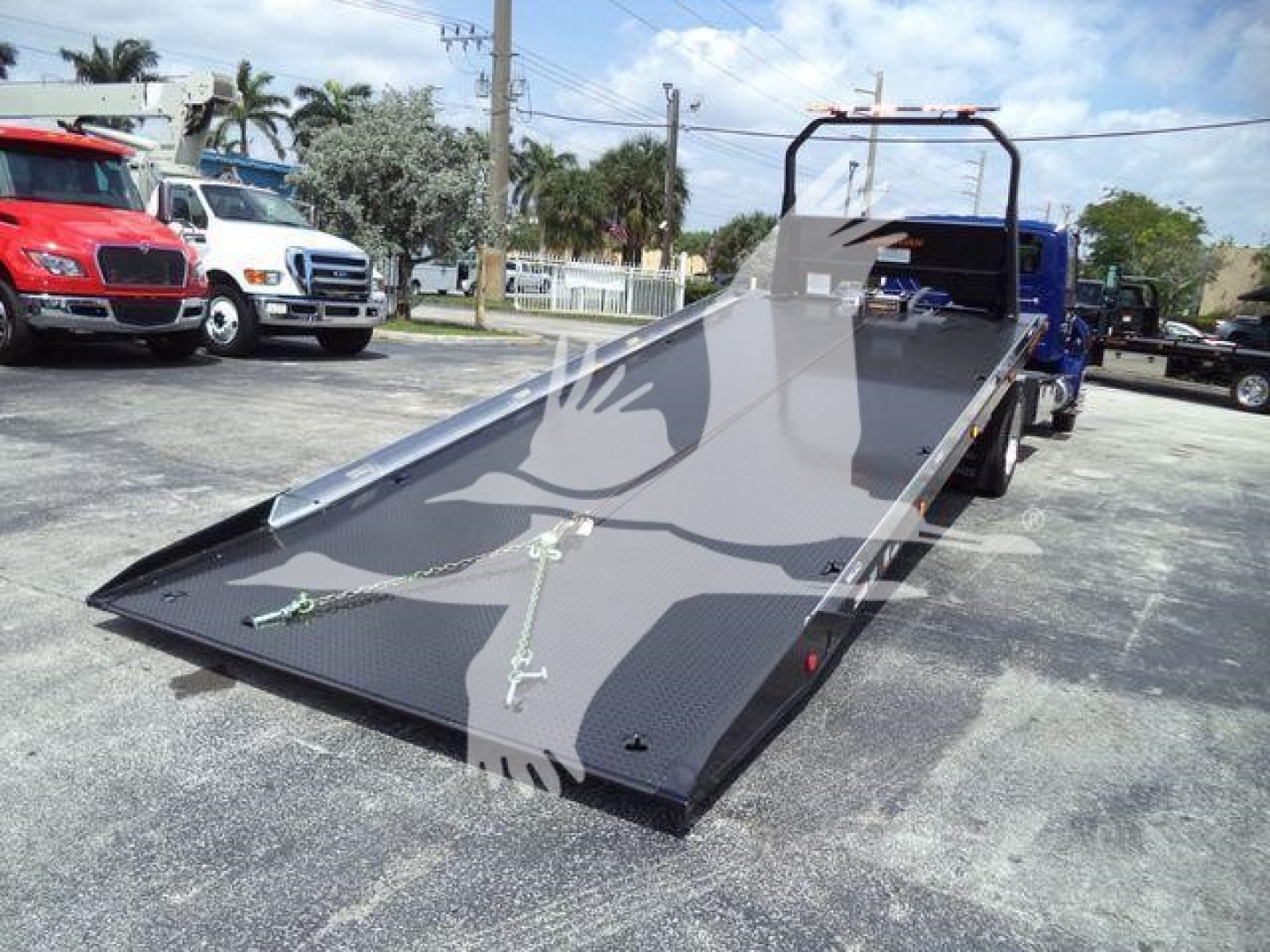 Flatbed tow truck with a hydraulic rollback bed in a loading position, showing metal ramps and chain tie-downs.