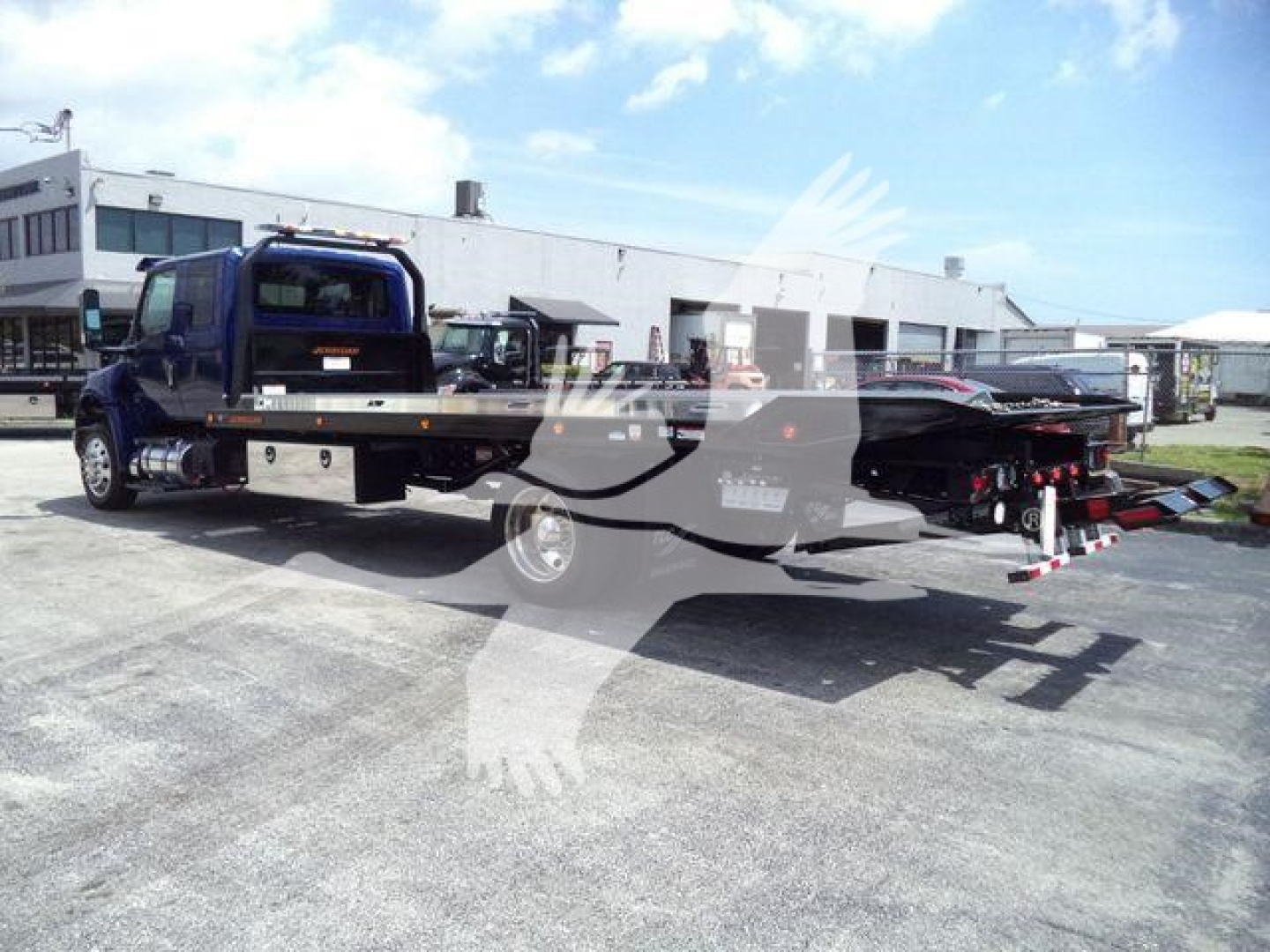 Truck flatbed tow truck with a blue cab and extended flatbed seen from the side in a lot.