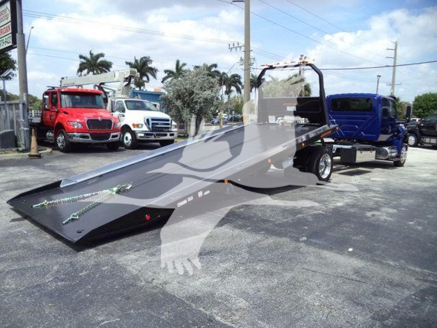 Truck tow rollback flatbed with a metal tilt bed and hydraulic ramp, at a dealership lot with multiple trucks in background.