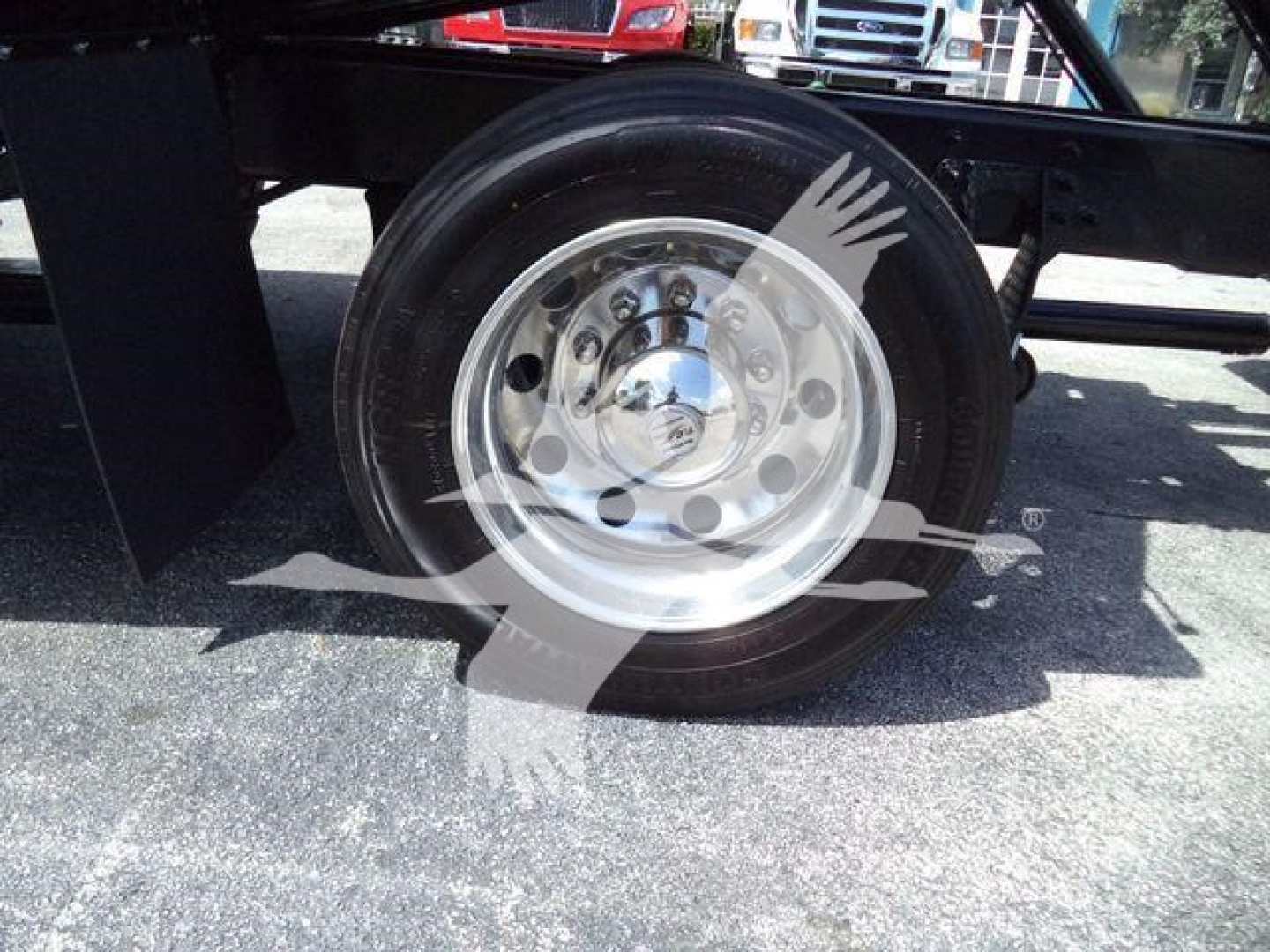 Wheel hub and tire of a truck trailer showing a polished metal hub with multiple lug nuts and a circular cover. The ground is asphalt.