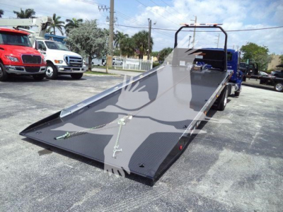Flatbed tow truck with a hydraulic rollback bed in a loading position, showing metal ramps and chain tie-downs.