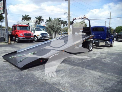 Truck tow rollback flatbed with a metal tilt bed and hydraulic ramp, at a dealership lot with multiple trucks in background.