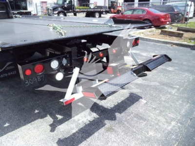 Truck rollback trailer rear with ramp and hydraulic controls showing metal platform, taillights, chains, and support legs.