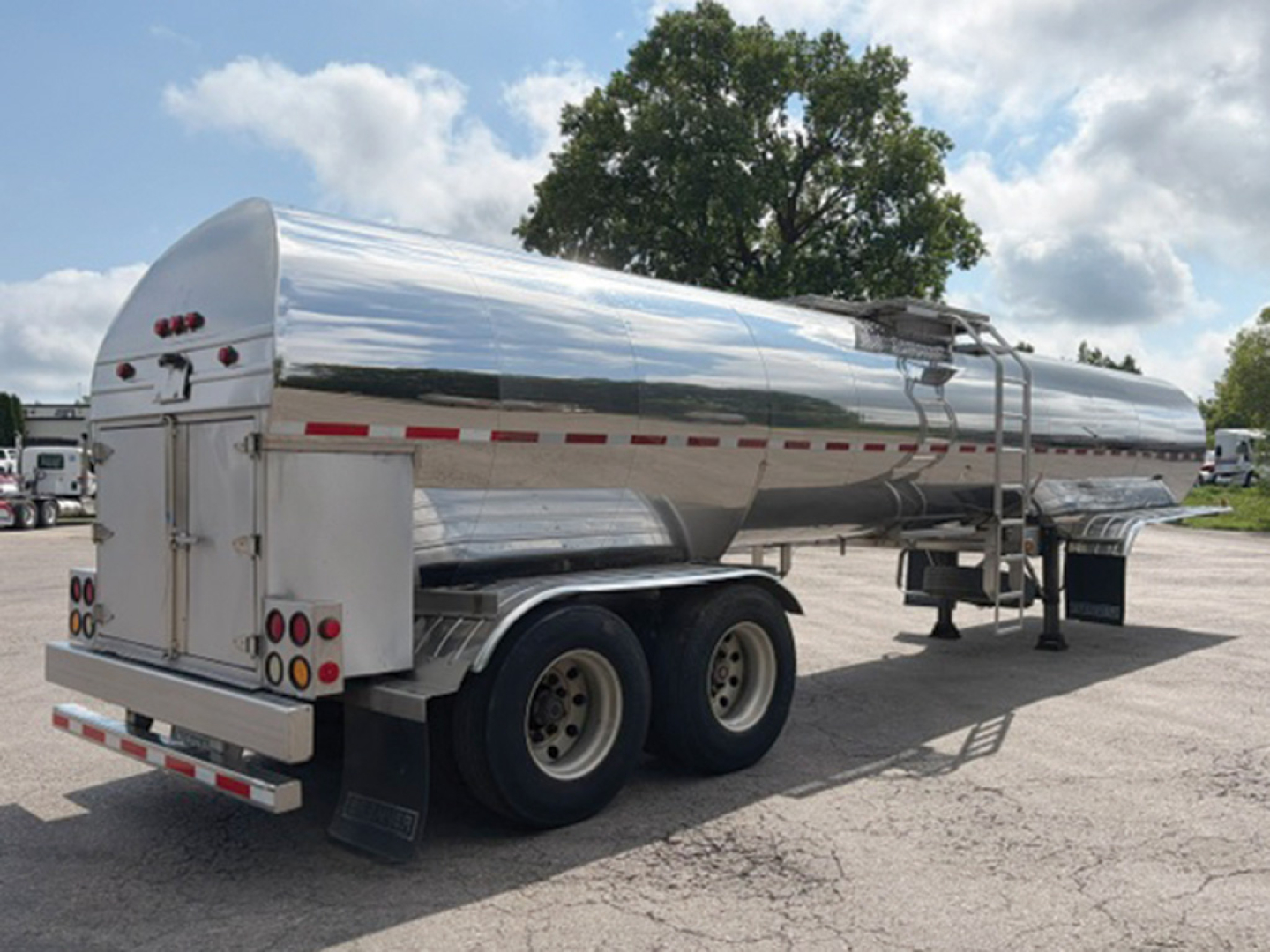 Tank trailer in side-rear view, polished chrome cylindrical tanker on a triple-axle chassis with rear doors and safety rails.