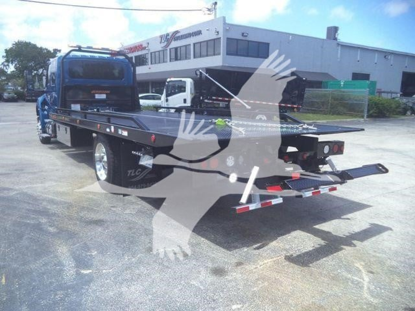 Flatbed rollback truck rear view showing long deck, hydraulic winches, and dual rear taillights; blue cab, loading ramps folded, in a dealership lot