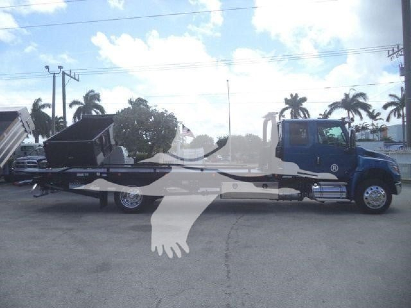 International MV rollback tow truck side view showing blue cab and long flatbed with a black wheel-lift vehicle on the bed; palm trees in background.