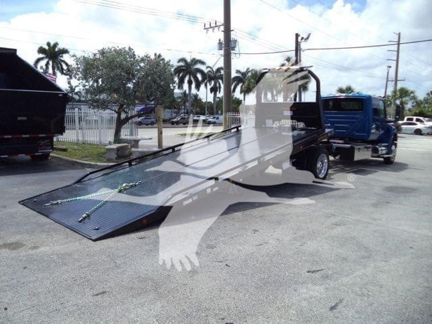 A flatbed rollback truck with a lowered ramp in a parking lot, blue truck in background, palm trees and power lines.