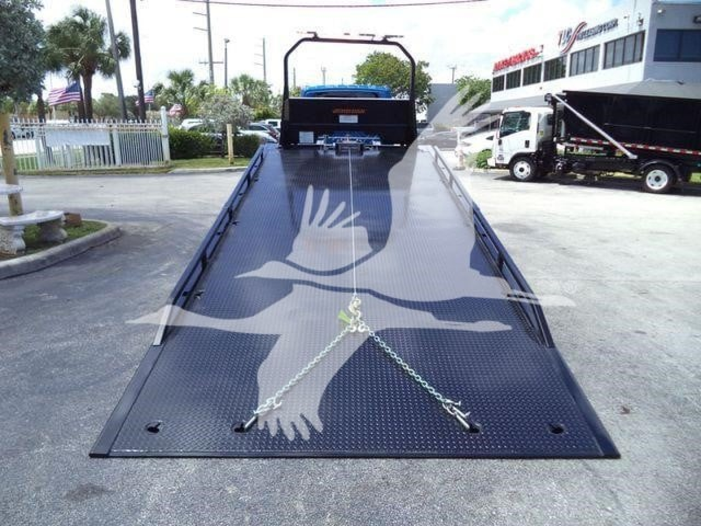 Mvable image showing a black roll-back tow truck ramp with chains, flatbed platform, and a two-tone vehicle in background.