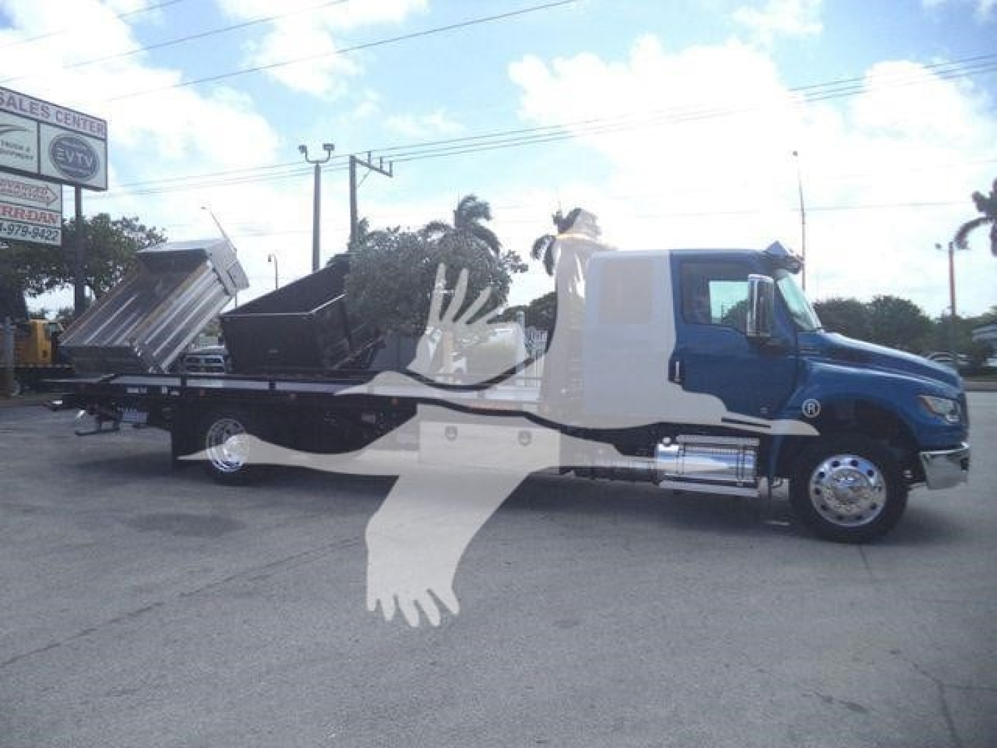 Truck in rollback configuration showing cab and flatbed with dump bed attached, blue cab, black bed, and hydraulic equipment visible.