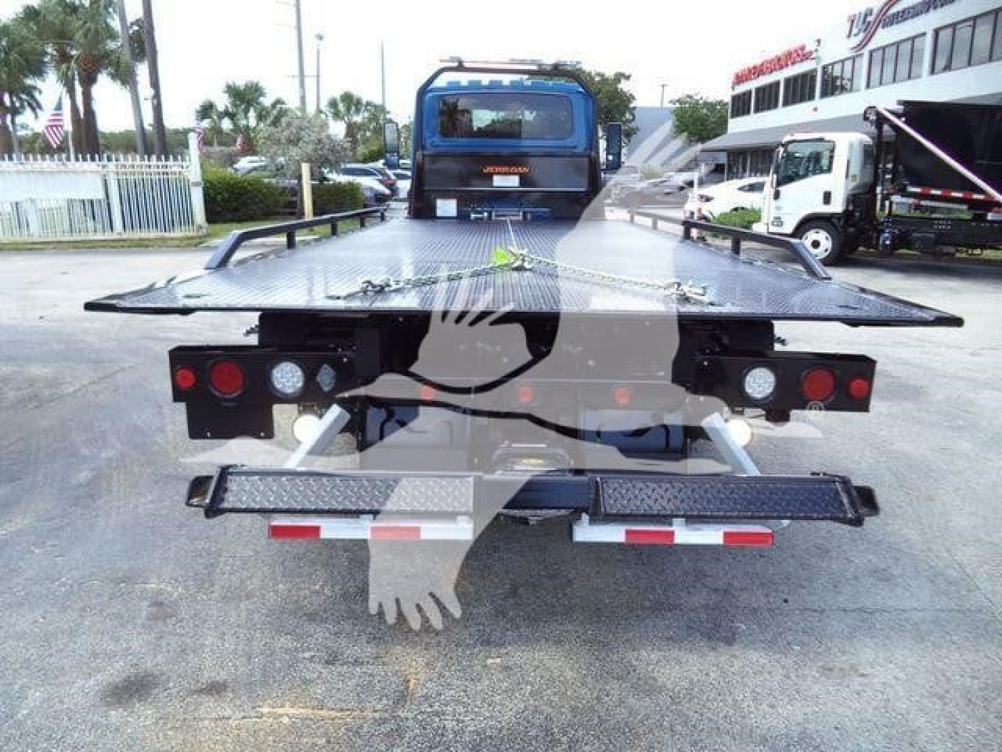 Tow truck rollback bed from rear showing flatbed with taillights, bumper, and hydraulic mechanism; International MV truck in background.