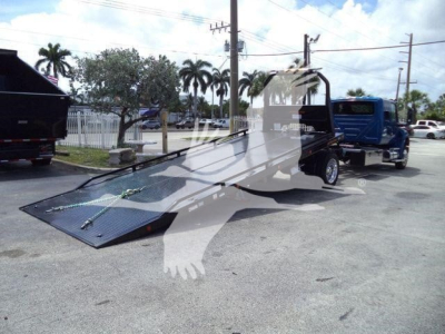A flatbed rollback truck with a lowered ramp in a parking lot, blue truck in background, palm trees and power lines.