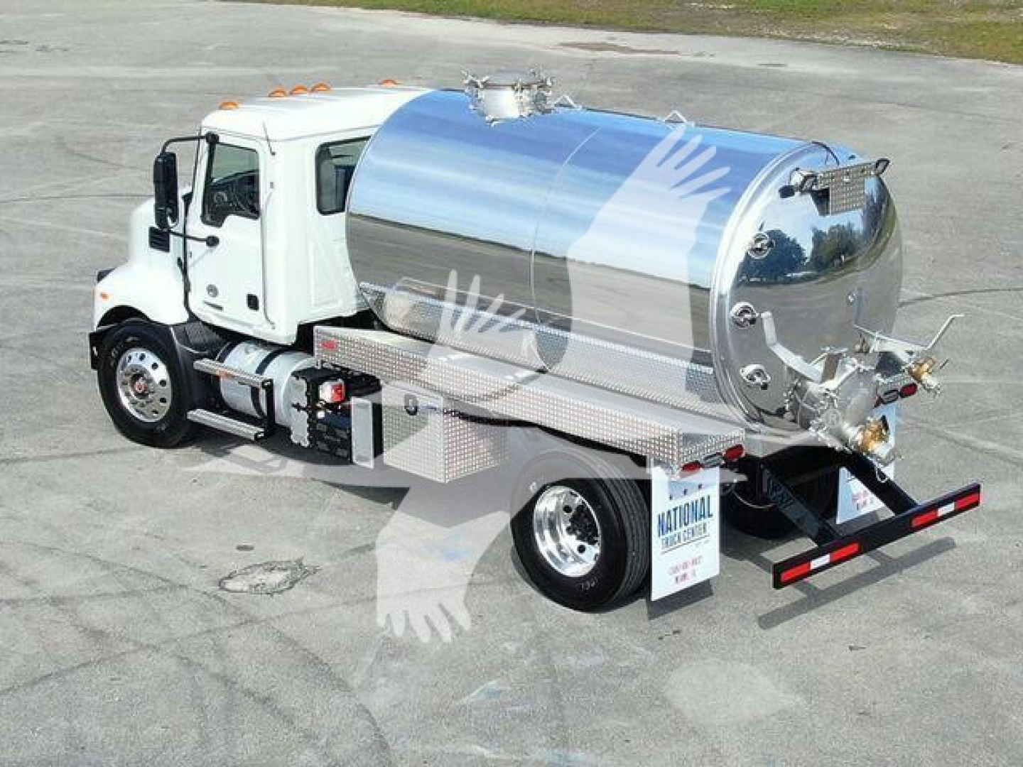 Truck with a polished stainless steel tank on a white cab. Side view shows rear hoses and plumbing fittings, mirrors, headlights, and a black bumper; tanker has circular manway and rear access panels.
