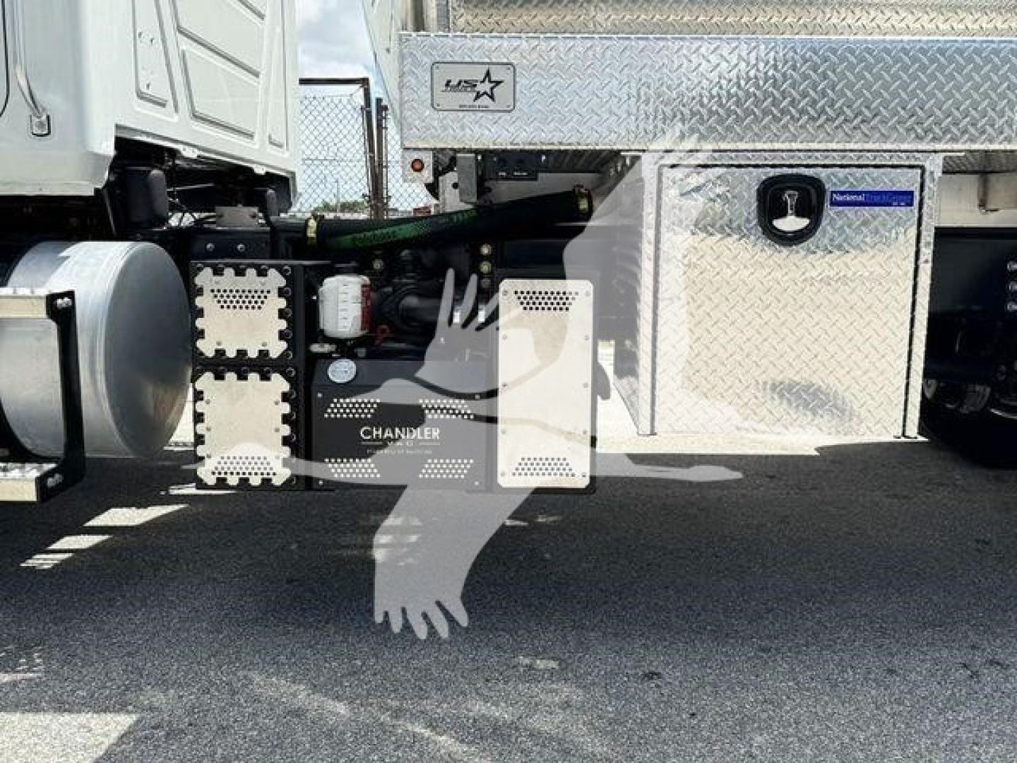 Truck undercarriage showing fuel tank and chunky aluminum toolbox with circular Mack badge and 'CHANDLER' label; components and hoses visible.