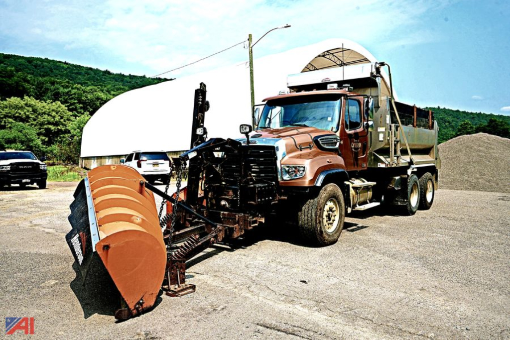 Truck view showing a brown Freightliner SD114 dump truck with attached snow plow and large front scoop, in a quarry setting.