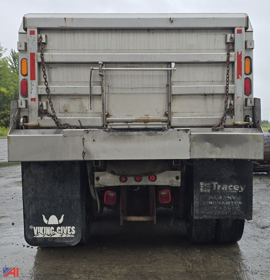 Dump truck rear showing spread tailgate, dual mud flaps, and chains with the hoist assembly; rear bumper and taillights visible, overcast day.