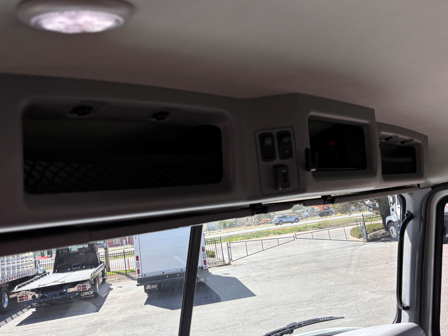 Interior view of a Mack GR64B truck cab area showing overhead compartments and switches; windshield and dashboard glimpses with exterior lot in background.