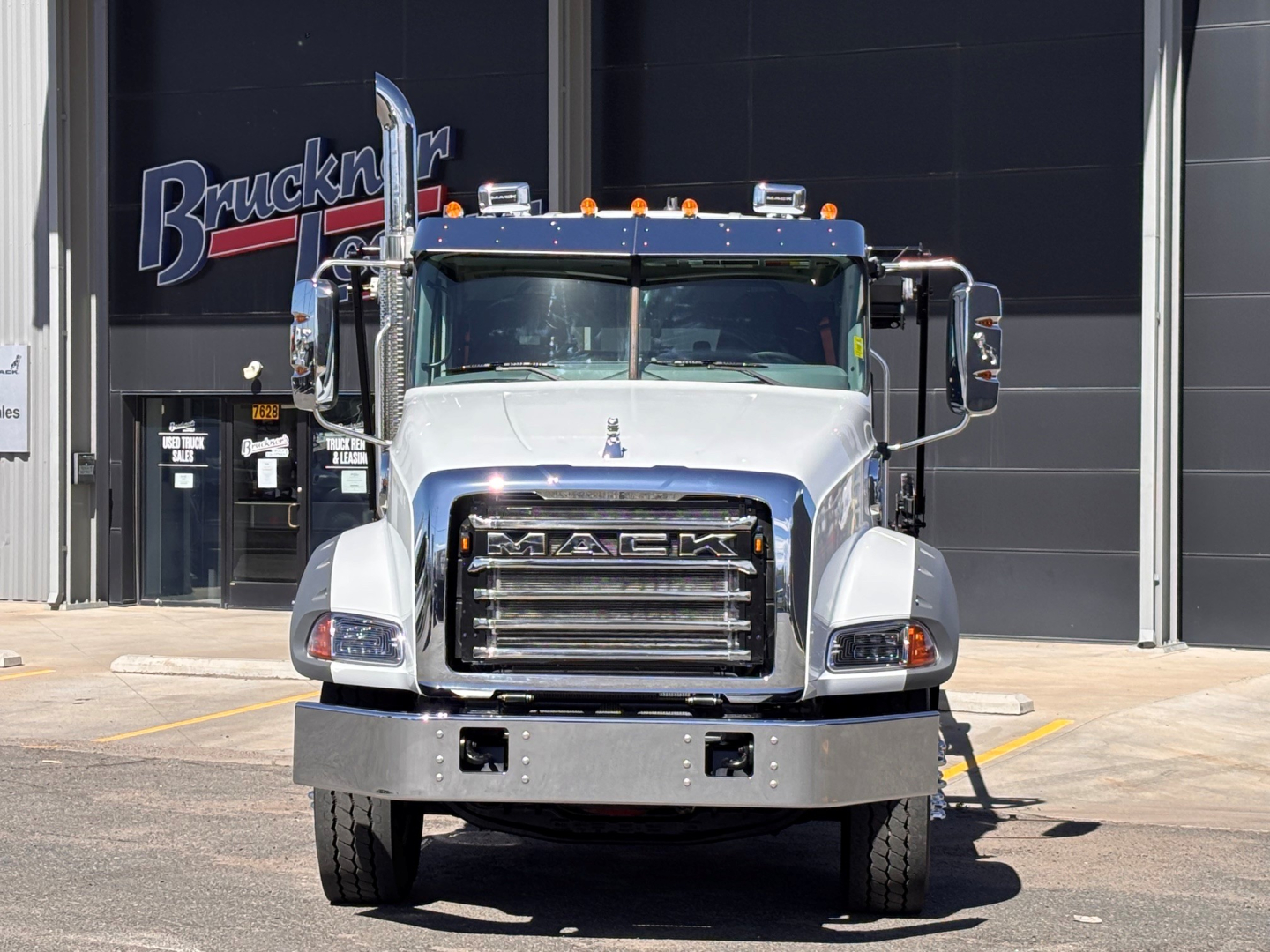 Front view of a white 2026 Mack GR64B roll-off truck with large grille, bumper, and side mirrors; dealership background with Brucken signage.