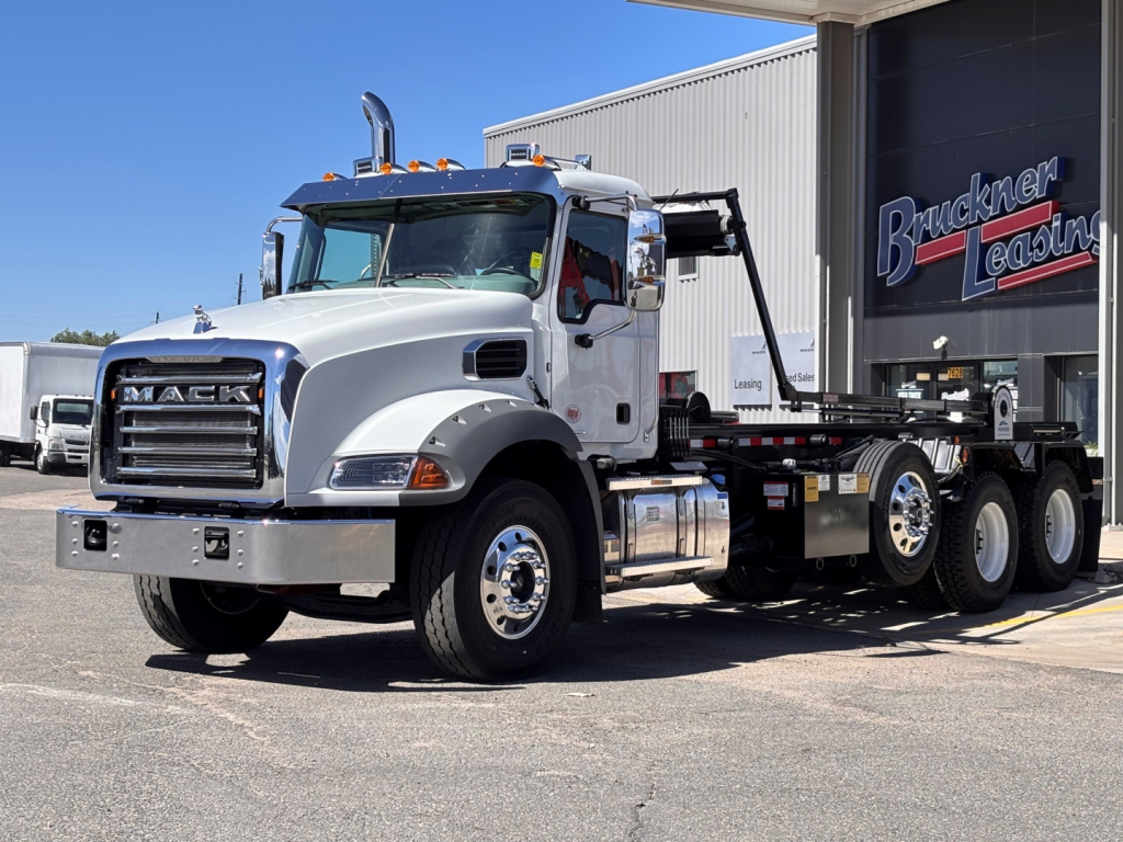 Front-left view of a white Mack GR64B rolloff truck with chrome grille, dual rear axles, and crew cab. Visible are Mack emblem, large front bumper, and a hook-lift on the flatbed.
