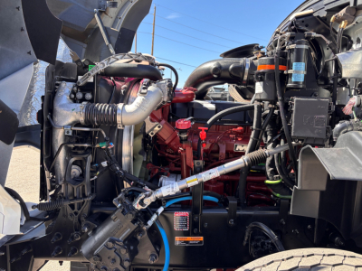 Mack GR64B engine bay showing red diesel engine, hoses, hoses clamps, and surrounding components in a truck cab. Visible labels and tubing across the open hood area.