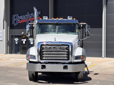 Front view of a white 2026 Mack GR64B roll-off truck with large grille, bumper, and side mirrors; dealership background with Brucken signage.