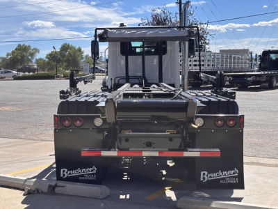Rear view of a new 2026 Mack GR64B roll-off truck showing rear bumper with taillights, mud flaps labeled 'Bruckner's Truck & Equipment' and wheel chocks, VIN and stock info in context.