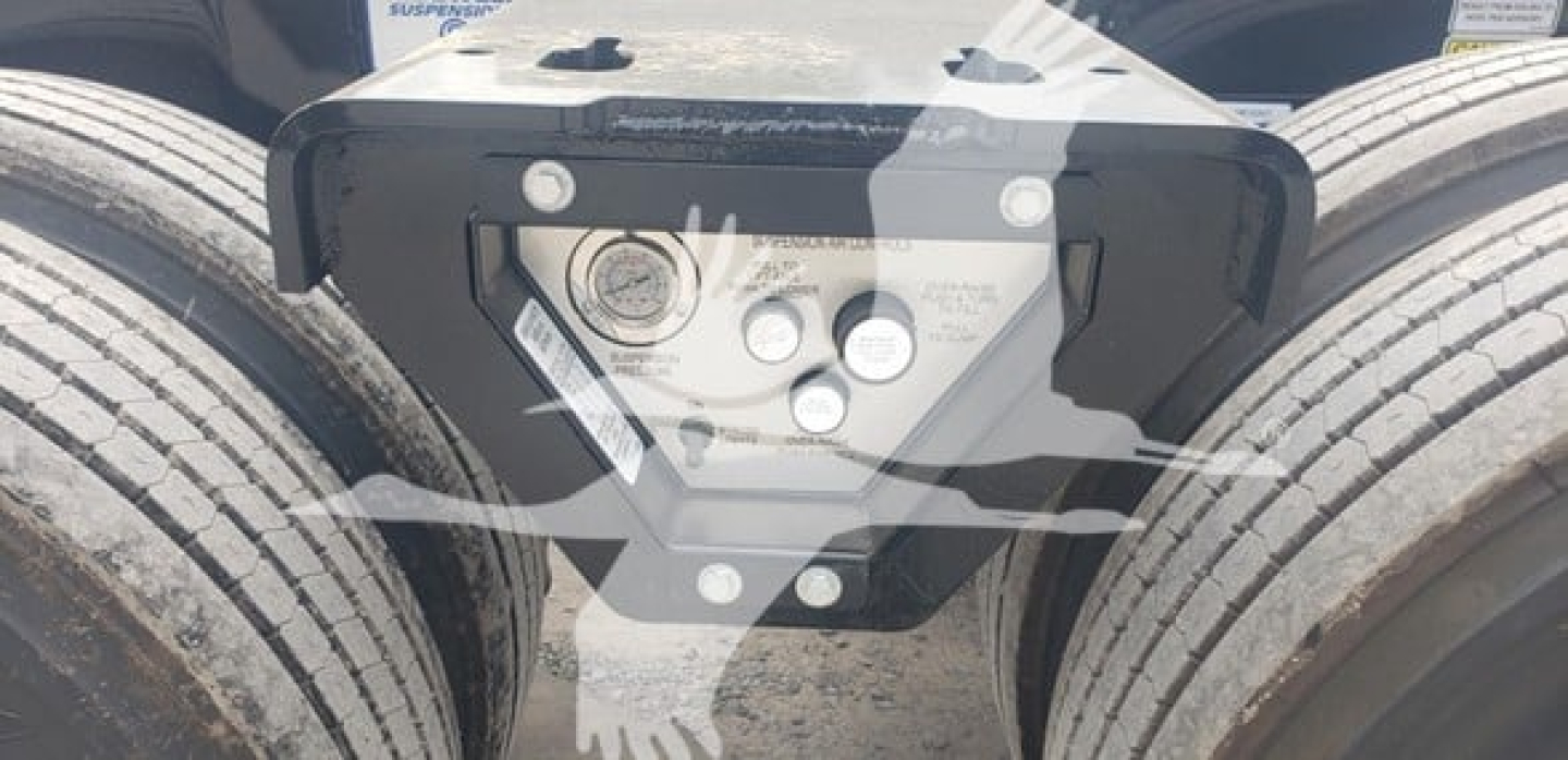 Trailer fifth wheel with mounting plate and control panel showing bolts and labels on black steel, two tires visible in background.