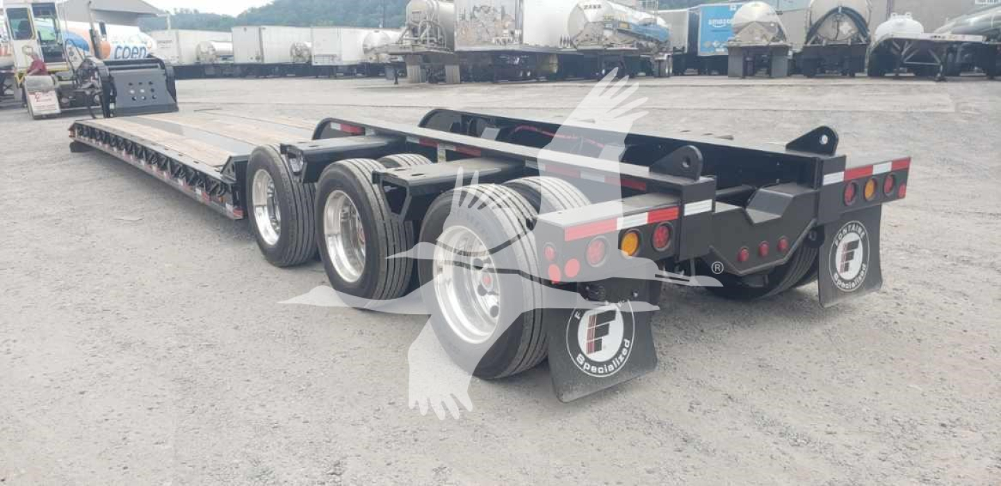 Trailer showing a Fontaine Magnitude 55Mx lowboy trailer with a long flatbed, triple-axle rear and front axle, black frame, red and white rear safety lights, and mudflaps.