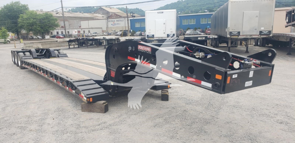 Lowboy trailer showing magnified gooseneck and deck with black frame and white graphics, on a gravel lot with other trailers in background.