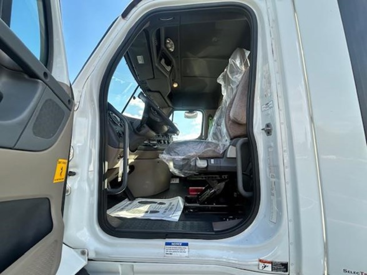 Cab interior of a Freightliner Cascadia truck with steering wheel, dashboard, driver seat, and plastic covers on seat; open door showing entry area and floor mat.
