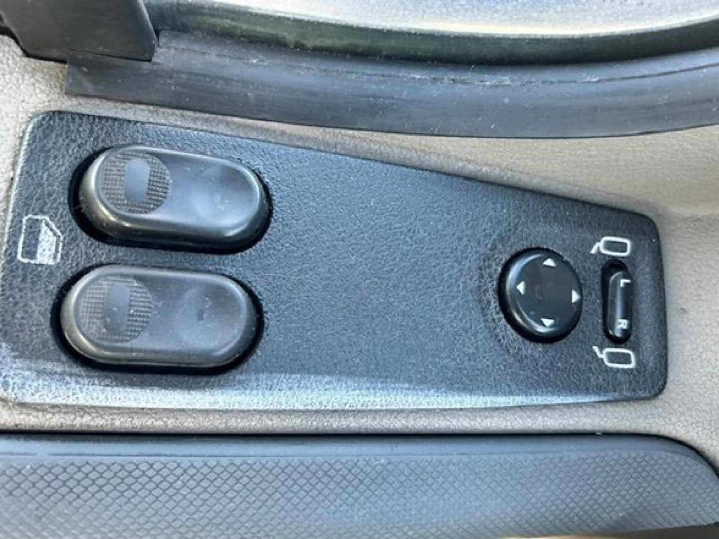 Truck interior switch panel showing two window/seat controls and a circular joystick-like control with arrows, plus a small cigarette lighter style outlet near the headlight icon.