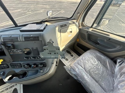 Cab interior of a Freightliner Cascadia showing steering wheel, dashboard, control panel, and a plastic-covered seat. Various panels and a mount bracket are visible.