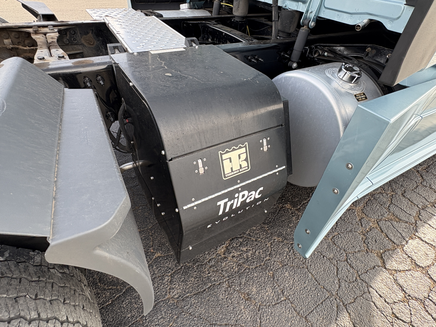 Tractor-trailer engine bay area showing Tripac auxiliary power unit (APU) with Triumph logo and a fuel tank nearby; part of Freightliner Cascadia truck.