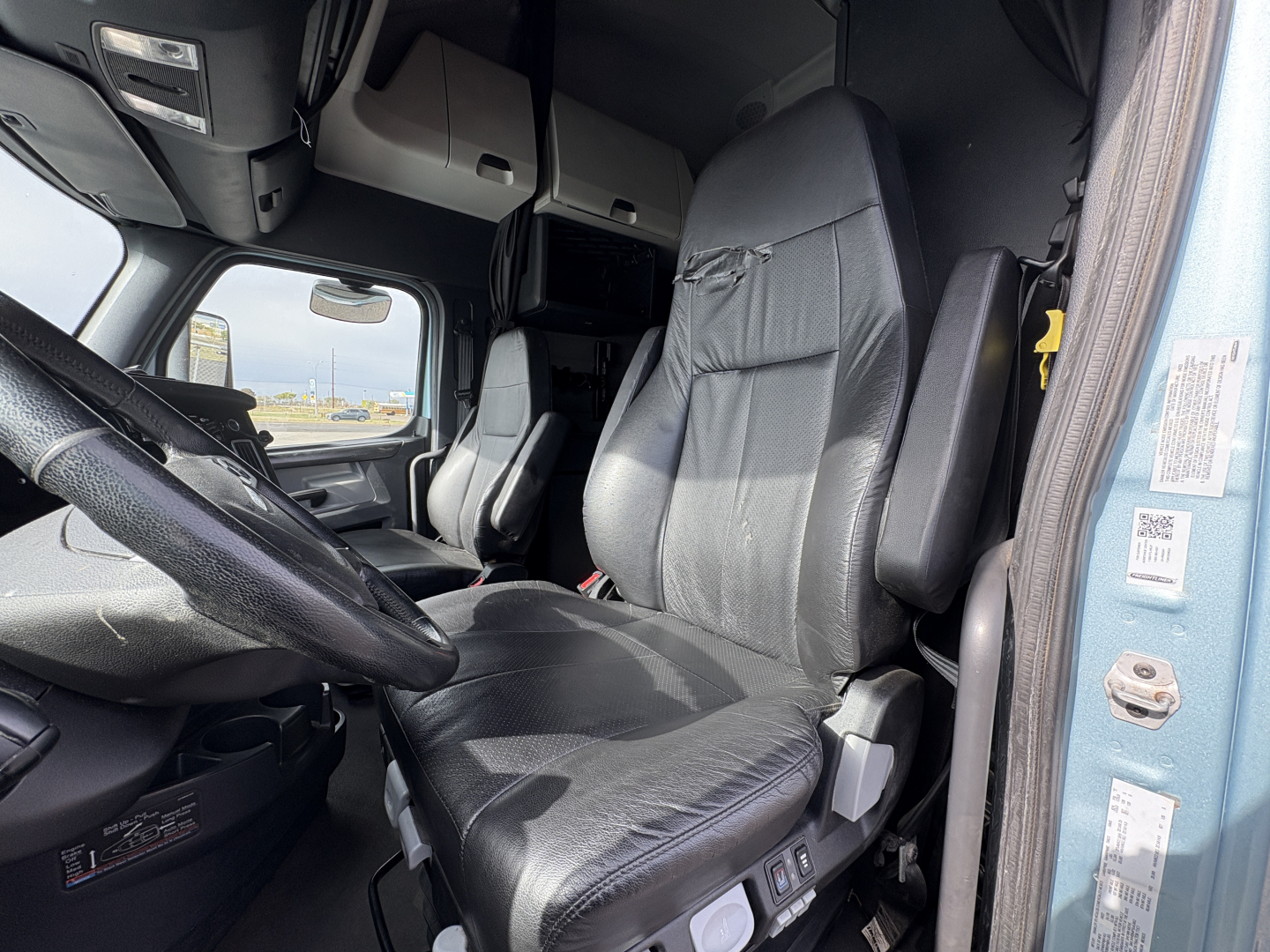Truck cab interior showing driver seat, steering wheel, dash, and overhead storage in a 2023 Freightliner Cascadia 126 (view from doorway).